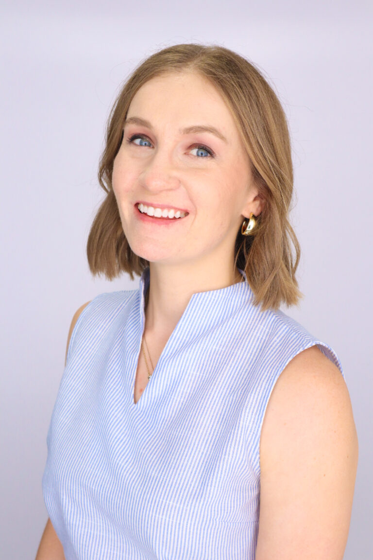 Katherine Norton smiles in a blue striped sleeveless top with gold hoop earrings.
