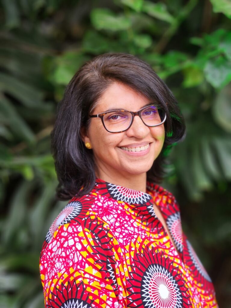 Sia Nowrojee smiles, wearing glasses and a colorful patterned shirt, against a green background.